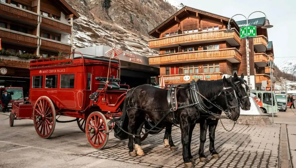 Vintage horse-drawn carriage in a mountain village with wooden buildings and snowy peaks in the back.